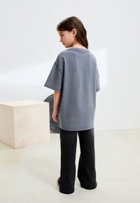 Niña con cabello castaño que lleva una camiseta gris holgada y pantalones anchos negros, de pie de lado sobre un suelo beige con decoración geométrica.