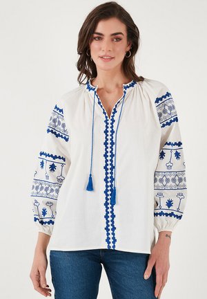 White blouse featuring blue embroidery, a tassel tie at the neckline, and patterned sleeves. Made of lightweight fabric, has a relaxed fit.