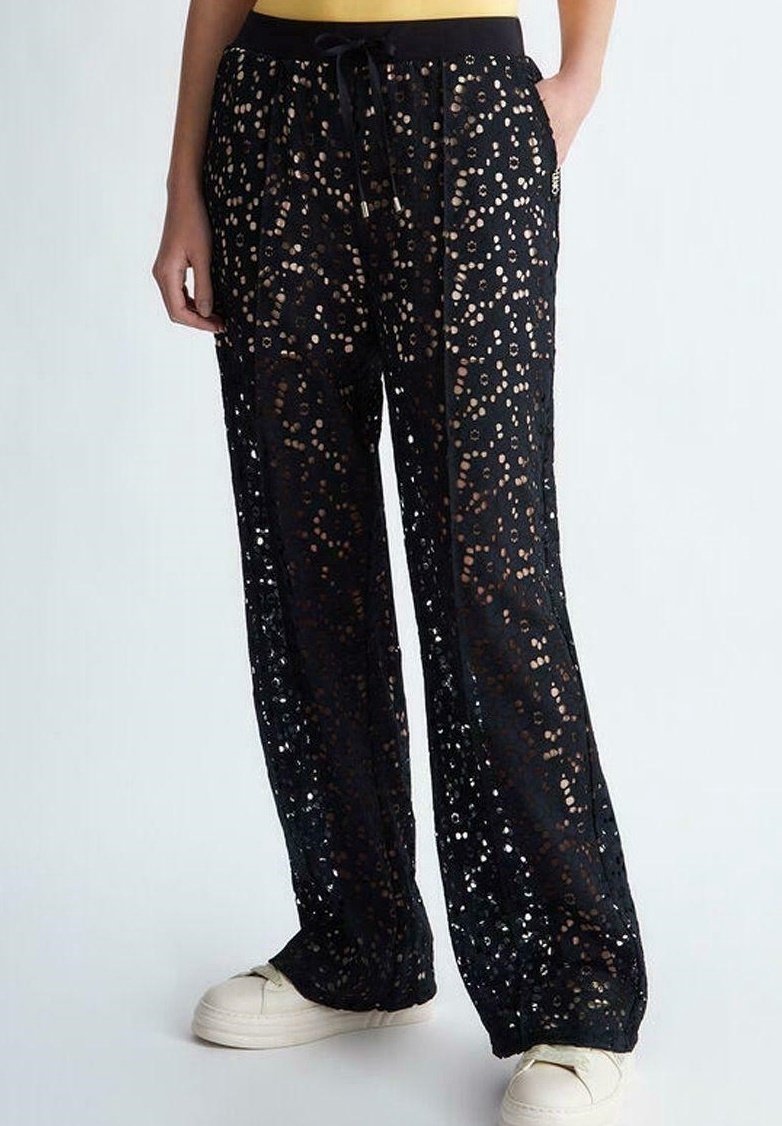 Pantalon en dentelle noire à coupe ample, taille élastique et motifs de petits pois métalliques dispersés. Porté avec des chaussures blanches.