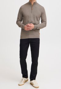 Maglione zip-up marrone chiaro con polsini e orlo a coste, abbinato a pantaloni neri e sneakers beige con strisce color miele.