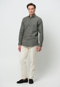 Grijs overhemd met een klassieke kraag, lange mouwen en zwarte knopen, gecombineerd met beige broek en donkere schoenen op een effen achtergrond.