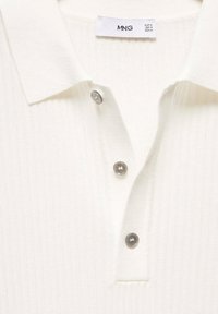 Polo blanc côtelé avec col, doté de trois boutons argentés et d'une étiquette de marque. Tissu texturé avec un motif vertical subtil.