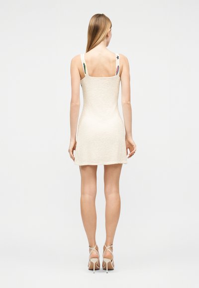 Γυναίκα με μακριά ξανθά μαλλιά που φοράει μια μίνι κρεμ αμάνικη φορέματα με κεντημένους ιμάντες και μπεζ ψηλοτάκουνα σανδάλια, στέκεται γυρισμένη προς τα πίσω.