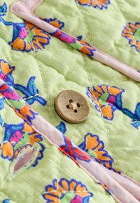 Tissu matelassé vert clair avec des motifs floraux vibrants en orange et bleu. Un bouton en bois à quatre trous est mis en évidence.