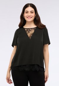 Donna con capelli scuri che indossa un top nero a maniche corte con dettagli in pizzo sul colletto e sull'orlo, sorridente contro uno sfondo semplice.