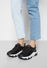 Sneaker sportive nere con tomaia in rete e pelle, suola intermedia bianca, dettagli dorati e suola testurizzata. Indossate con jeans azzurri chiari.