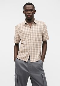 Πουκάμισο με κοντά μανίκια σε καρό σχέδιο σε ανοιχτό καφέ και μπεζ, με κουμπιά και χαλαρή εφαρμογή. Συνδυάζεται με γκρι παντελόνι cargo με τσέπες.