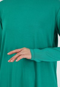 Chemise turquoise à manches longues en tissu doux, avec une coupe décontractée et une texture subtile. Détails aux poignets visibles.