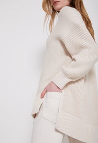 Žena v krémovém pleteném svetru s dlouhými rukávy a krémových kalhotách, s rukou v kapsách kalhot, na jednobarevném pozadí.