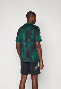 Μπλουζάκι κοντό μανίκι πράσινης παραλλαγής από αναπνεύσιμο υλικό με μαύρες λεπτομέρειες και λευκές ρίγες στους μανίκους, σε συνδυασμό με μαύρες βερμούδες.