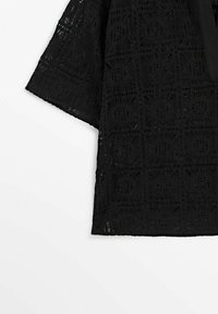 Haut noir à manches mi-longues avec motif en dentelle crochetée en carrés sur un fond uni clair.