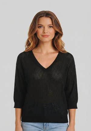 Femme aux cheveux ondulés portant un pull à col en V en maille noire transparente à manches trois-quarts et un jean bleu clair, devant un fond uni.