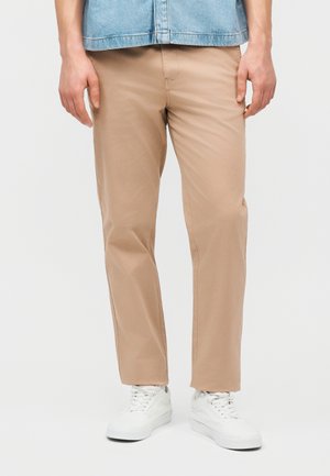 Κάτω σώμα ατόμου που φοράει μπεζ chinos, λευκά αθλητικά παπούτσια και ένα ανοιχτό μπλε τζιν πουκάμισο, στέκεται μπροστά σε απλό φόντο.