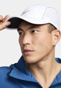 Hvid Nike-kasket med en buet skygge, der har et gråt logo og teksturerede sidepaneler, båret lidt til siden. Blå jakke synlig nedenunder.