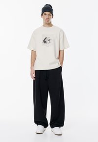 T-shirt oversize color crema con stampa grafica in bianco e nero, abbinata a pantaloni neri larghi e sneakers bianche, completata da un berretto nero.