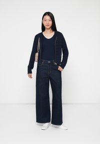 Granatowy sweter zapinany na zamek na granatowej bluzce, w połączeniu z szerokimi ciemnymi dżinsami i białymi sneakersami. Na ramieniu noszona jest mała jasna torba.