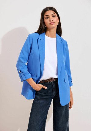 Femeie cu păr lung și închis la culoare, purtând un sacou albastru aprins peste o cămașă albă și blugi închiși la culoare, stând cu o mână în buzunar pe un fundal simplu.