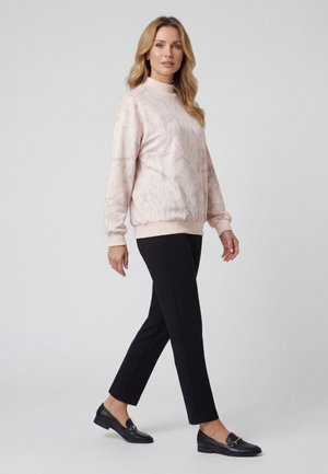 Donna che cammina indossando una felpa rosa chiaro con fantasia, pantaloni neri corti e mocassini neri su uno sfondo grigio chiaro.