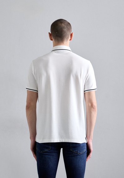 White polo shirt with contrast black trim, short sleeves, soft texture, and a straight hem. View from the back, paired with blue jeans.