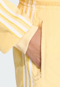 Žltý teplákový súprava z teri látky s bielymi a žltými pruhmi na rukávoch. Obsahuje vrecko so zipsom. Mäkká textura a uvoľnený strih.