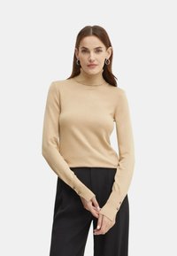 Maglione beige a collo alto realizzato in tessuto morbido, con maniche lunghe con dettagli in bottone sui polsini e una silhouette aderente.