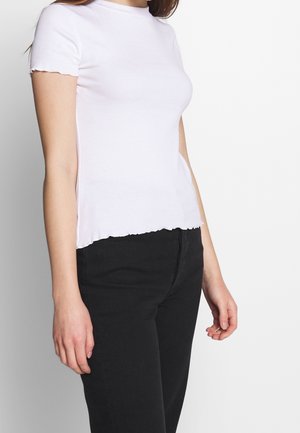 Woman wearing a fitted white short-sleeve ribbed top with lettuce hem and black high-waist pants, standing against a plain background.