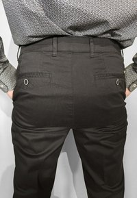 Persona che indossa pantaloni grigio scuro con trama e una camicia a maniche lunghe con motivo, mani nelle tasche anteriori, vista dalla schiena dalla vita in giù.