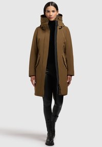Manteau imperméable marron avec un col haut, une capuche et des zips, présentant des poches latérales et une coupe ajustée. Porté sur des leggings noirs et des bottes.