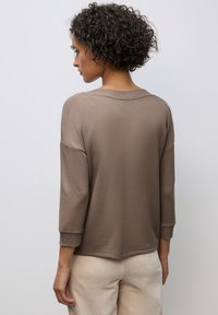 Brauner Pullover aus Jerseystoff mit lockerer Passform, 3/4-Ärmeln, geripptem Rundhalsausschnitt und Bündchen sowie glatter Textur; kombiniert mit hellbeigen Hosen.