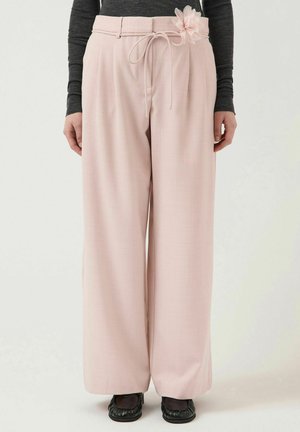 Kvinde iført højtaljede, vidde ben lyserøde bukser med snøre og blomsterdetalje, kombineret med sorte sko og en mørk bluse med lange ærmer.