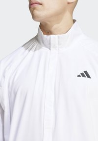 Witte sportjack met een hoge kraag, met een gladde textuur en een zwart logo op de borst. Minimalistisch ontwerp zonder patronen.