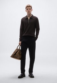 Ruda megzta megztinis su užtrauktuku, juodos kelnės ir juodi batai. Modelis laiko rudą odinę rankinę su kišenėle ir tvirtais rankenomis.