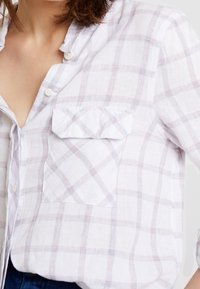 Chemise blanche à carreaux en tissu léger. Elle présente un col boutonné et une poche avec un rabat plié, dans un motif de grille rose et bleu.