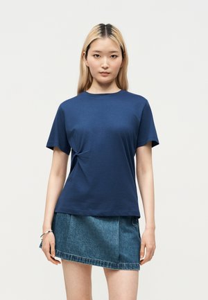 Jonge vrouw met steil blond haar draagt een marineblauw T-shirt met korte mouwen en een asymmetrische blauwe spijkerrok, staand tegen een effen achtergrond.