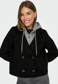 Mlada žena se smiješeći nosi crni dvoredni kaput preko sivog dukserice s patentnim zatvaračem i sivi hlače.