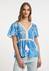 Blusa blu a fantasia con maniche corte, scollo a V, bordo in pizzo e vita con coulisse caratterizzata da un dettaglio con nappe bianche. Indossata con jeans chiari.