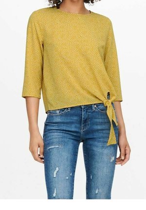 Femme portant un haut jaune à motifs avec manches 3/4, noué sur le côté, et un jean bleu déchiré.