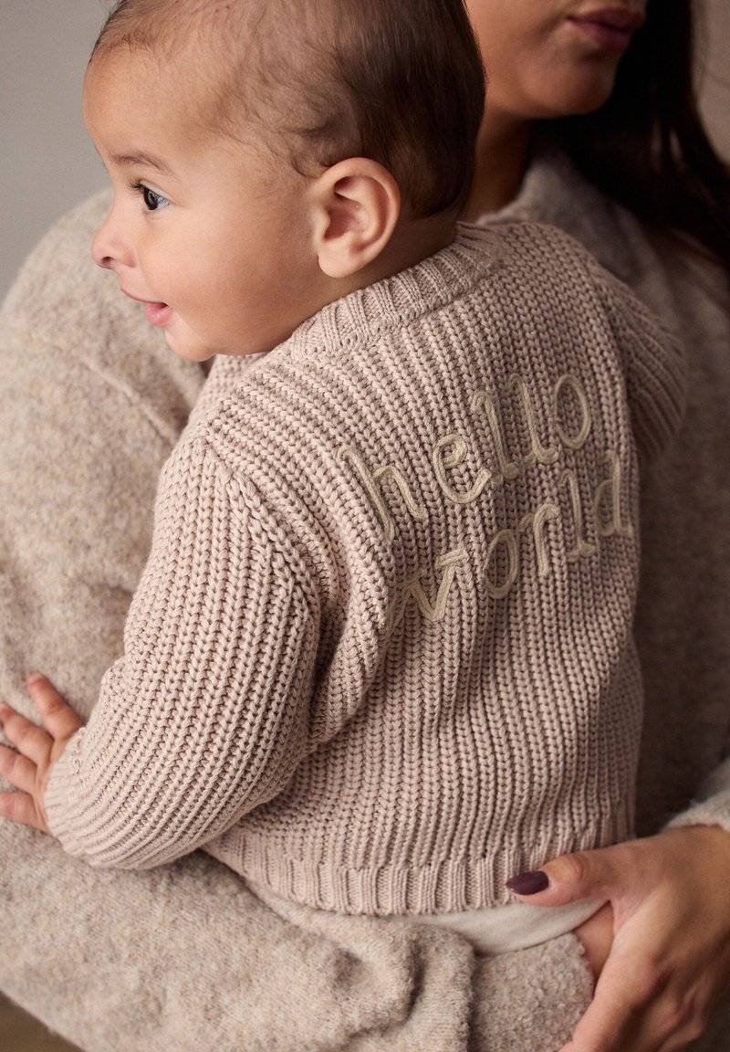 Cardigan en tricot de couleur beige doux avec le texte "hello world" brodé en vert clair au dos. Le tissu présente un motif texturé et côtelé.
