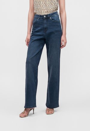 Personne portant un jean taille haute bleu foncé à jambes larges, associé à des sandales à talons hauts ouvertes et un top sans manches à motifs.