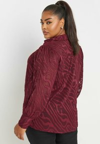Blusa bordò a maniche lunghe realizzata in tessuto testurizzato con un leggero motivo ondulato, dotata di colletto e polsini abbottonati.