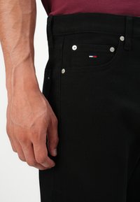 Nærbilde av en hånd som hviler ved siden av svarte denimjeans med sølvnitter og et lite rødt, hvitt og blått brodert flagg på lommen.