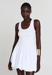 Femme à la peau foncée portant une robe sans manches blanche et un large bracelet en or martelé à son bras droit, sur un fond uni.