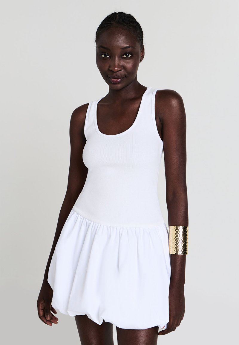 Femme à la peau foncée portant une robe sans manches blanche et un large bracelet en or martelé à son bras droit, sur un fond uni.