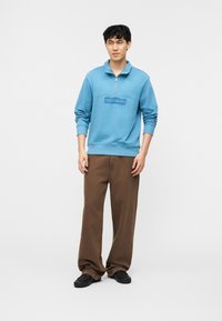 Νεαρός άνδρας στέκεται φορώντας μπλε φούτερ με φερμουάρ στο λαιμό, καφέ παντελόνι φαρδύ στο κάτω μέρος και μαύρα παπούτσια, μπροστά σε απλό λευκό φόντο.