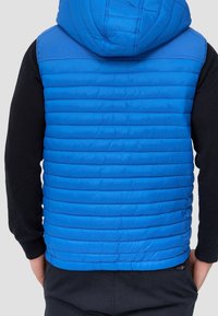 Chaleco acolchado azul con capucha, con secciones verticales acolchadas, combinado con una camiseta de manga larga negra y pantalones oscuros.