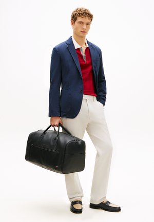 Ung mann i marineblå blazer, rød poloshirt, hvite bukser og svarte loafers holder en stor svart lærreiseveske.