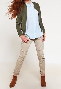 Olivově zelená bomber bunda přes světle modrou košili s knoflíky, spárovaná s béžovými kalhotami a hnědými kotníkovými botami. Hladké textury a pohodlný střih.