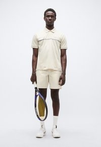 Ανοιχτό μπεζ πόλο πουκάμισο με γκρι οριζόντια λωρίδα, συνδυασμένο με μπεζ σορτς. Λευκά αθλητικά παπούτσια και ρακέτα τένις στο χέρι· ομαλή υφή σε όλο το σύνολο.