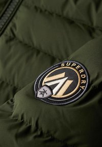 Olijfgroene pufferjack-stof met een gevoerd textuur. Bevat een ronde, zwart en gouden geborduurde badge met "Superdry."