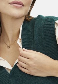 Teal gebreide trui met een diepe V-hals. Gelaagd over een witte blouse met kraag. Gouden hartvormige ketting. Gladde huid en natuurlijke handgebaren.